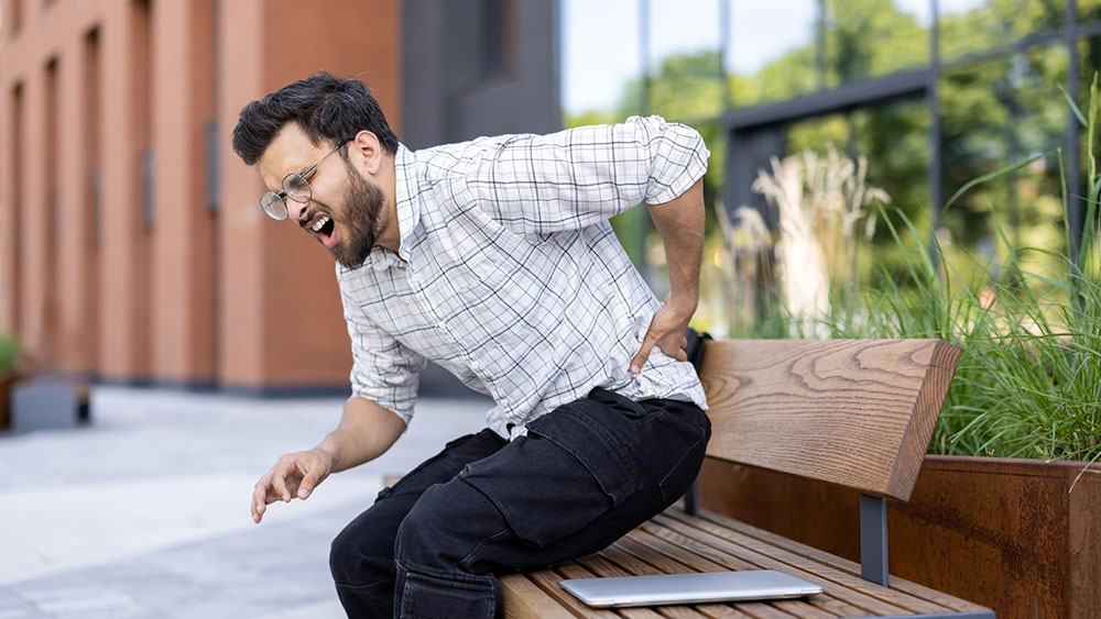 A young man on a bench holds his back, suffering from sciatica and in great pain